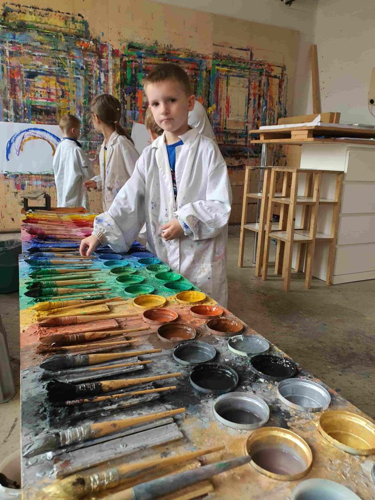 Ein Junge im Malerkittel schaut in die Kamera während er sich für einen Pinsel und Farbe entscheidet.