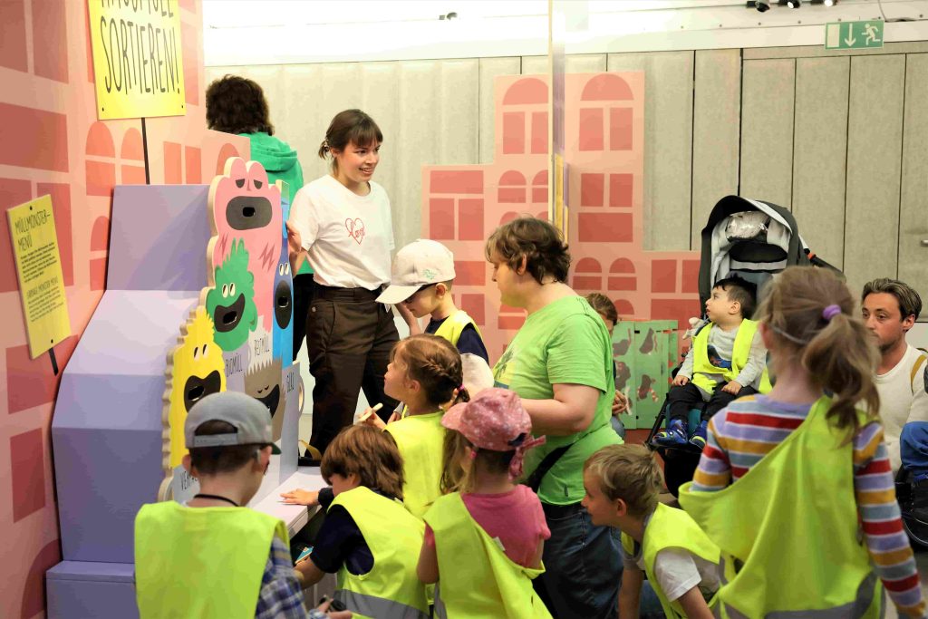 Eine Erwachsene zeigt einer Gruppe von Kindern und Erwachsenen ein Mülltrennungsspiel.